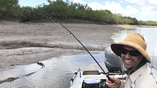 Lee grinning under a straw hat in a boat, rod bent, while a crocodile swims alongside