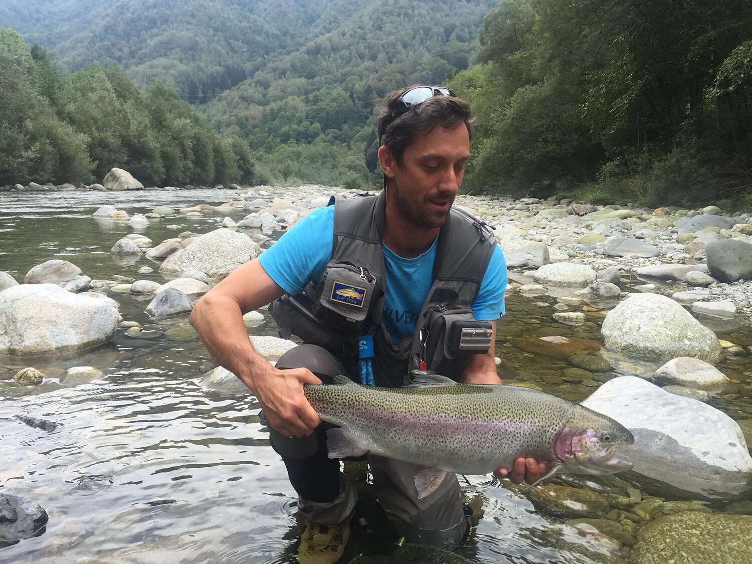 Lee crouching in a clear, rocky alpine river, holding a large rainbow trout